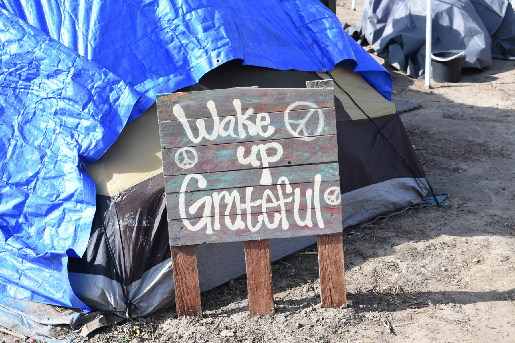 A tent with a tarp over it with a sign in front with stylized text reading "Wake up Grateful"