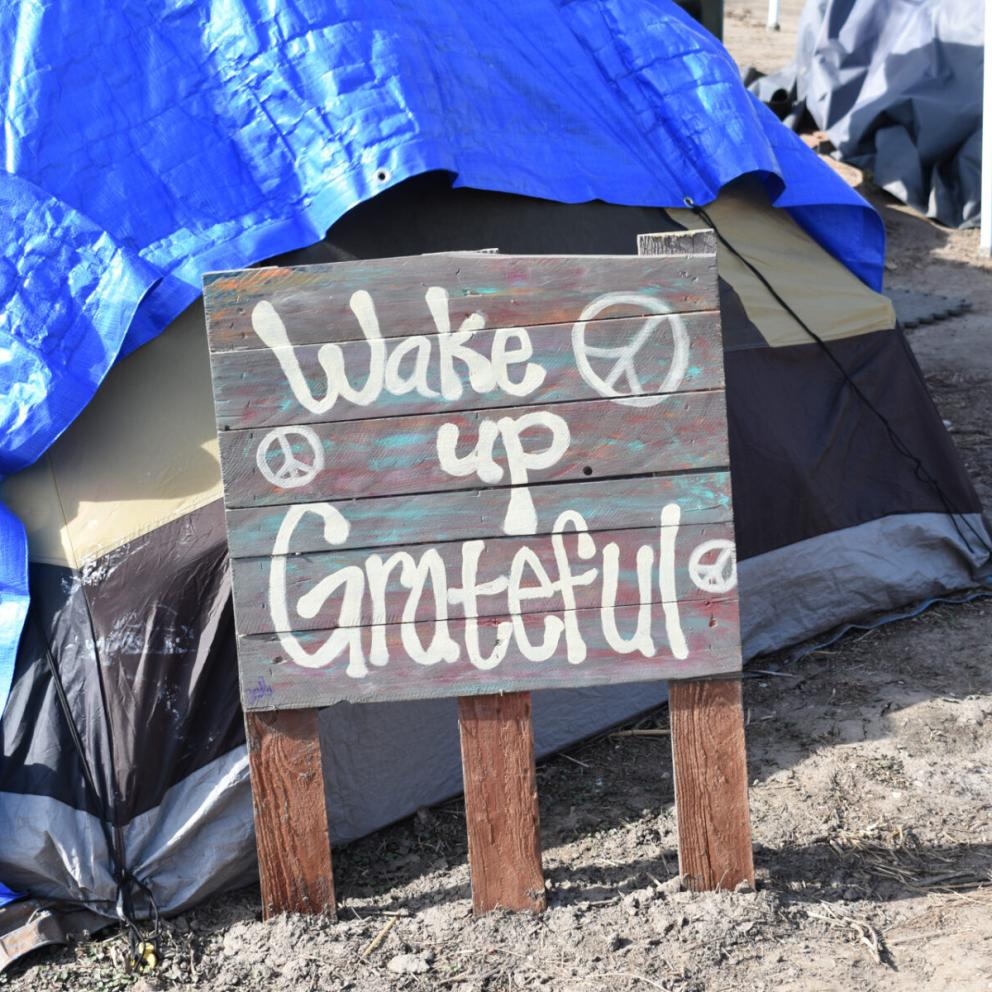 A tent with a tarp over it with a sign in front with stylized text reading "Wake up Grateful"
